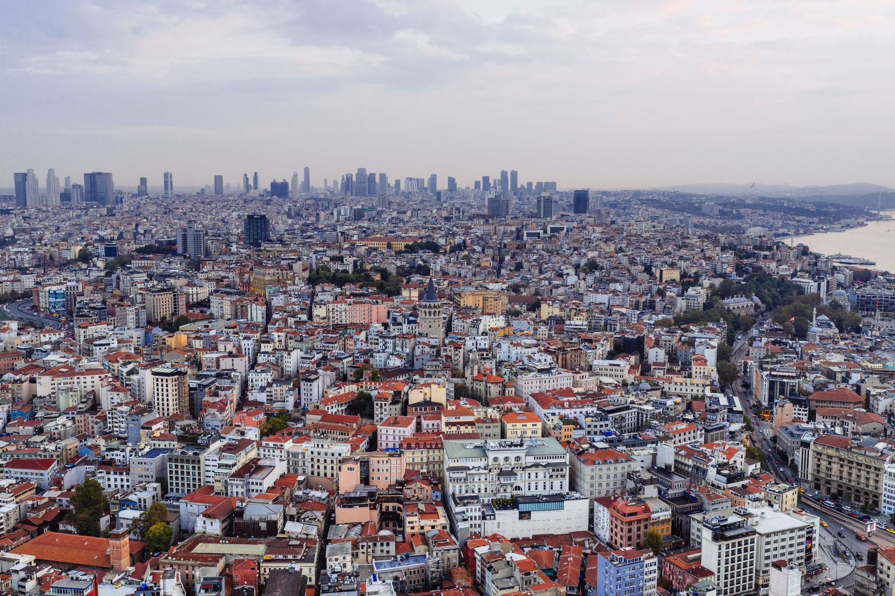 Aerial view over the dense rooftops of Istanbul toward a distant high-rise skyline under a soft evening sky