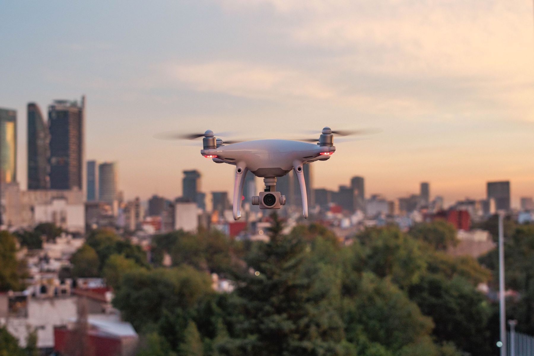 White quadcopter drone hovering above trees with a city skyline behind it at dusk