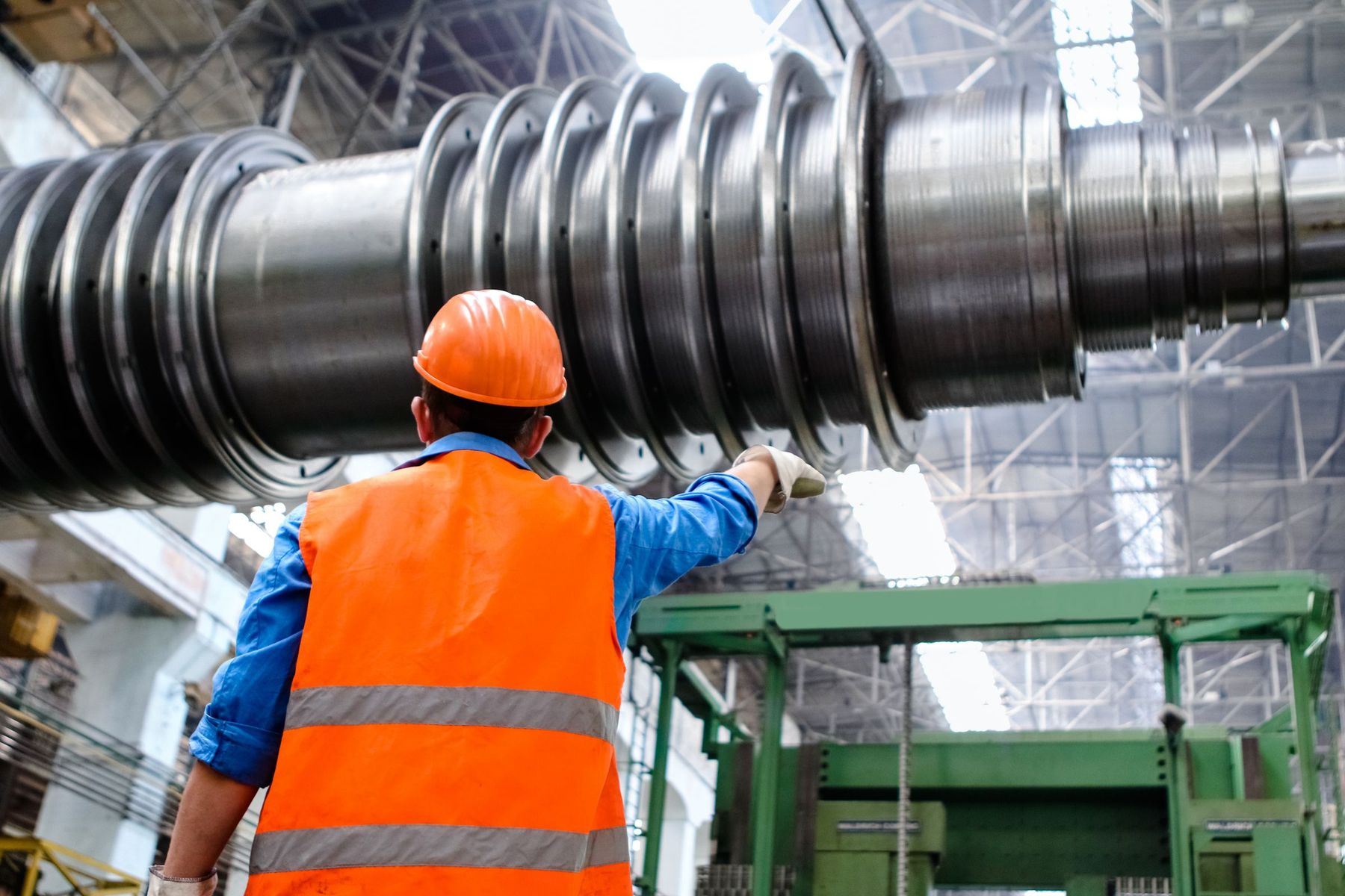 Engineer in an orange hard hat and hi-vis vest, seen from behind, inspecting a large turbine rotor shaft in a factory hall